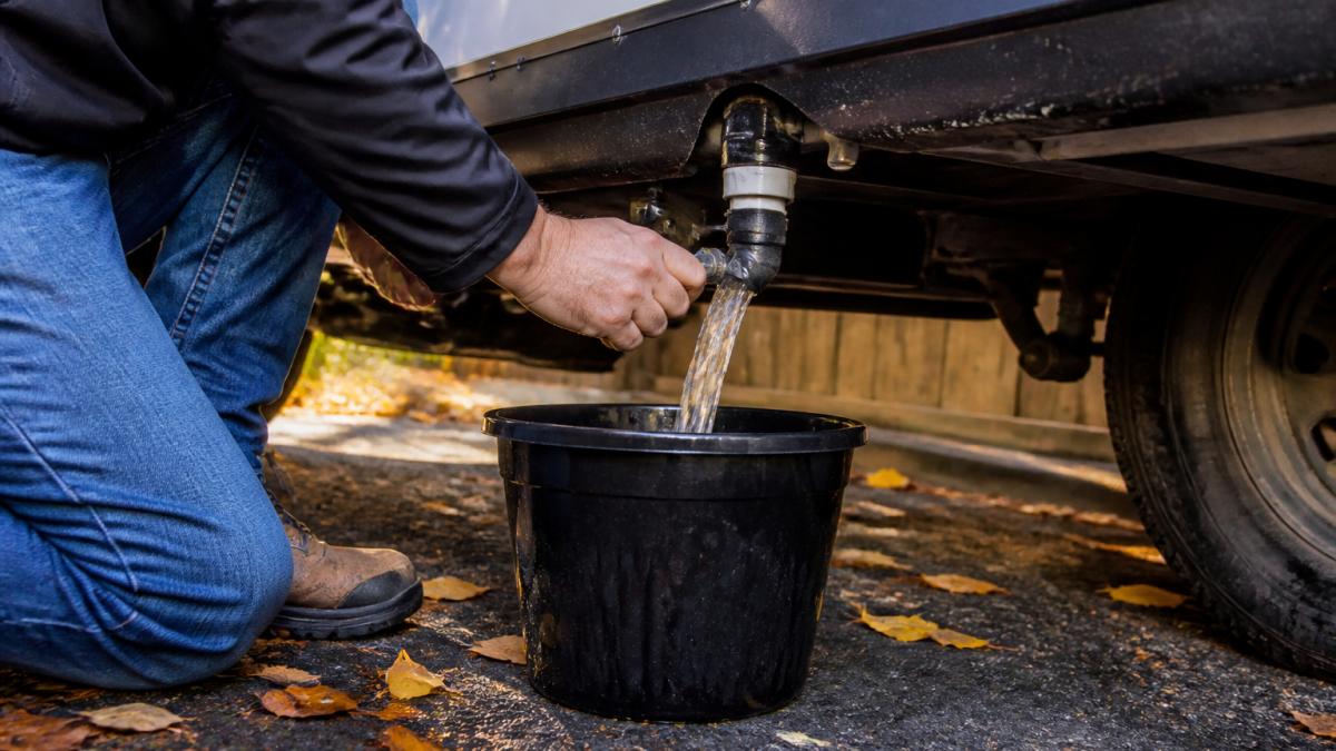 Close-up of hands opening a fresh water tank drain valve on an RV, with a bucket catching water underneath, parked in a driveway with autumn leaves nearby