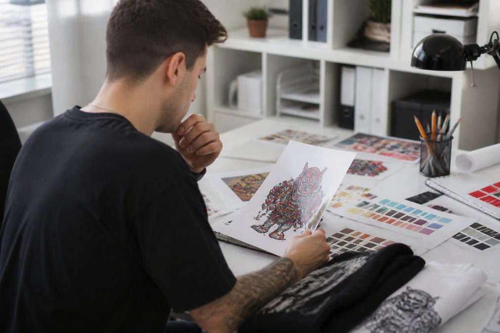 Designer reviewing artwork on colour-calibrated computer monitor with print-ready specifications displayed, checking design details before submission in professional office with reference materials and colour swatches visible
