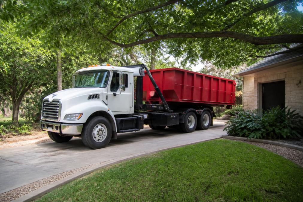 A roll-off dumpster delivery truck backing into a residential driveway with a clear view of the overhead clearance between the truck cab and low-hanging tree branches