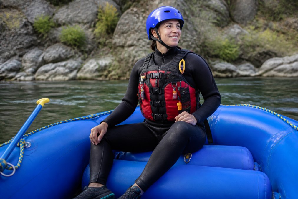 Professional rafter wearing proper whitewater gear including fitted wetsuit, Coast Guard-approved life jacket with multiple attachment points, protective helmet with chin strap, and water shoes with good grip, sitting confidently in a raft on calm water before entering rapids