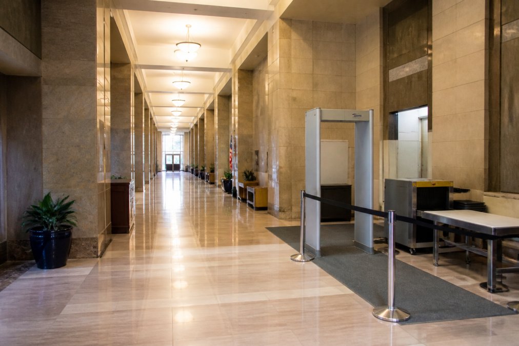 Interior of a modern courthouse hallway with professional lighting, security checkpoint area with metal detectors, directory signs on walls, and formal architectural elements conveying the serious legal environment