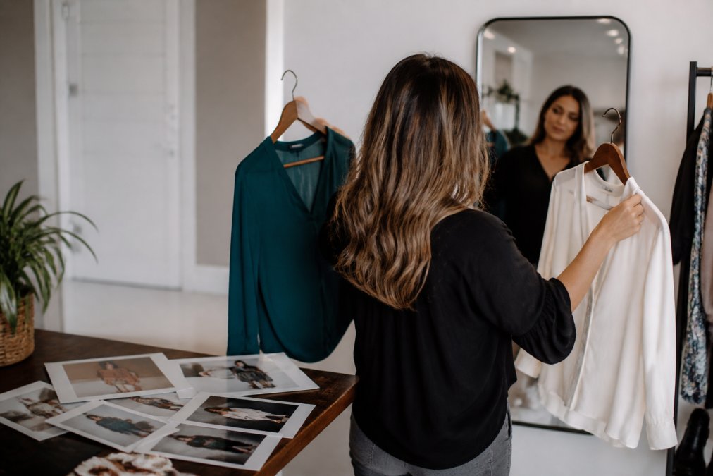 A person holding two solid-colored tops on hangers in front of a mirror, comparing options for a professional photoshoot, with a mood board of reference images visible on a table nearby
