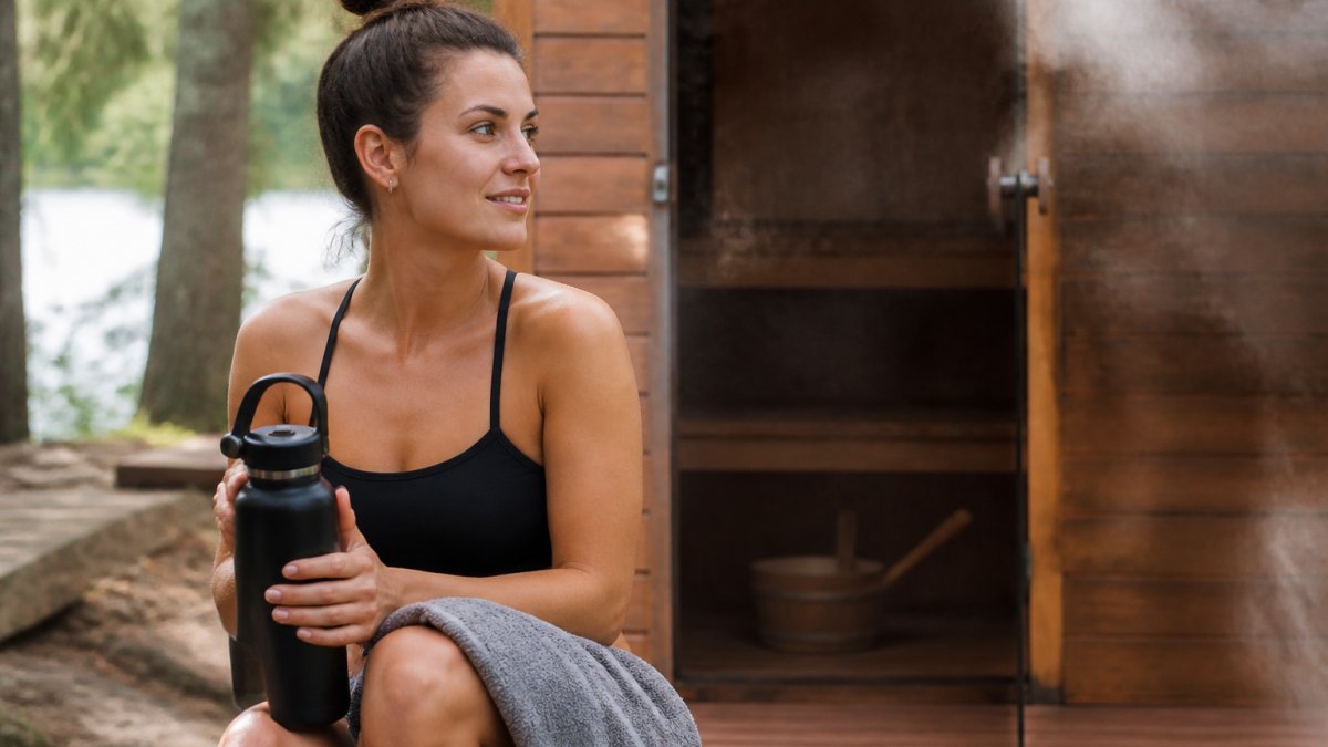 Person holding a water bottle and towel, sitting near a wooden sauna with steam rising in the background