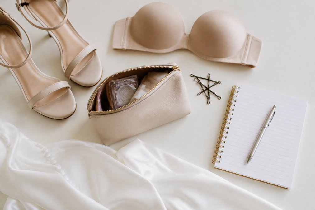 Flat-lay photograph on a clean white surface showing wedding fitting essentials: nude-coloured heels, a strapless bra, a small clutch bag with hair pins, and a notepad with a pen, shot in bright natural window light