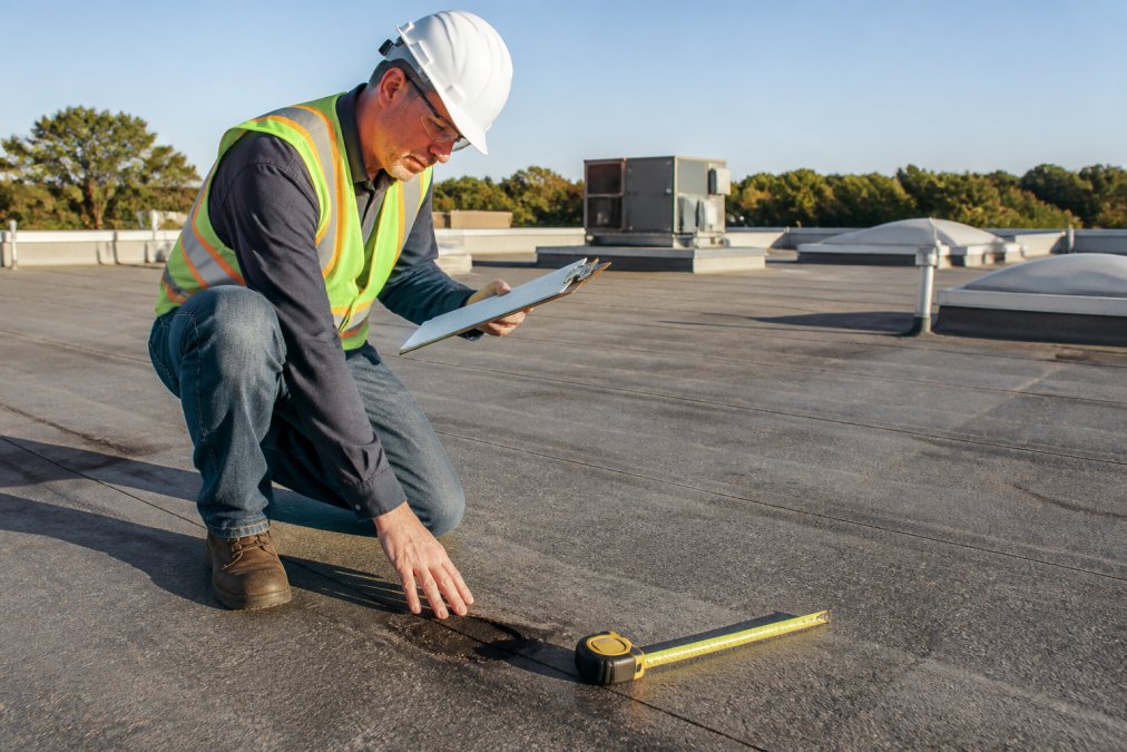 Professional roofer in safety equipment inspecting a commercial roof deck, checking for water intrusion and structural damage with measuring tools and clipboard visible in morning sunlight