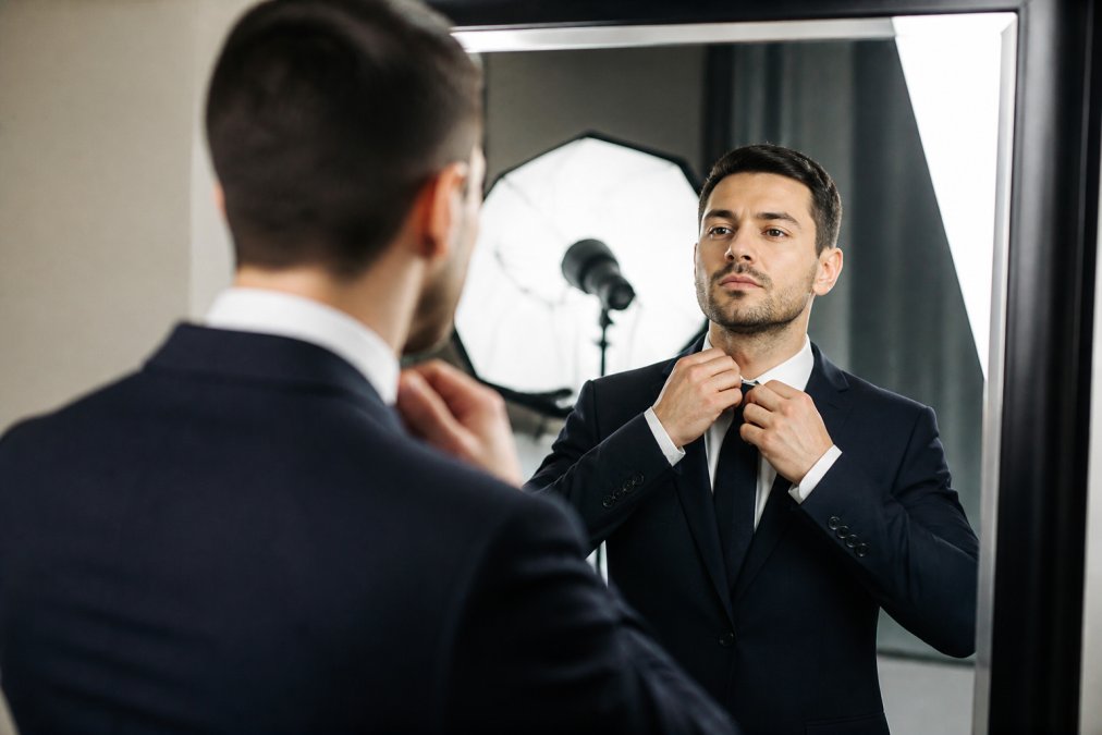 Professional man in a tailored suit adjusting his collar while checking his reflection in a mirror, bright studio lighting with softbox reflections visible
