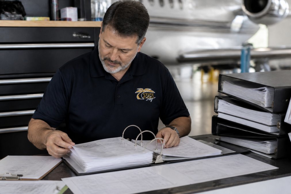 Aircraft owner or maintenance technician reviewing organized maintenance logbooks and documentation at a desk with files, binders, and technical records spread out under bright office lighting