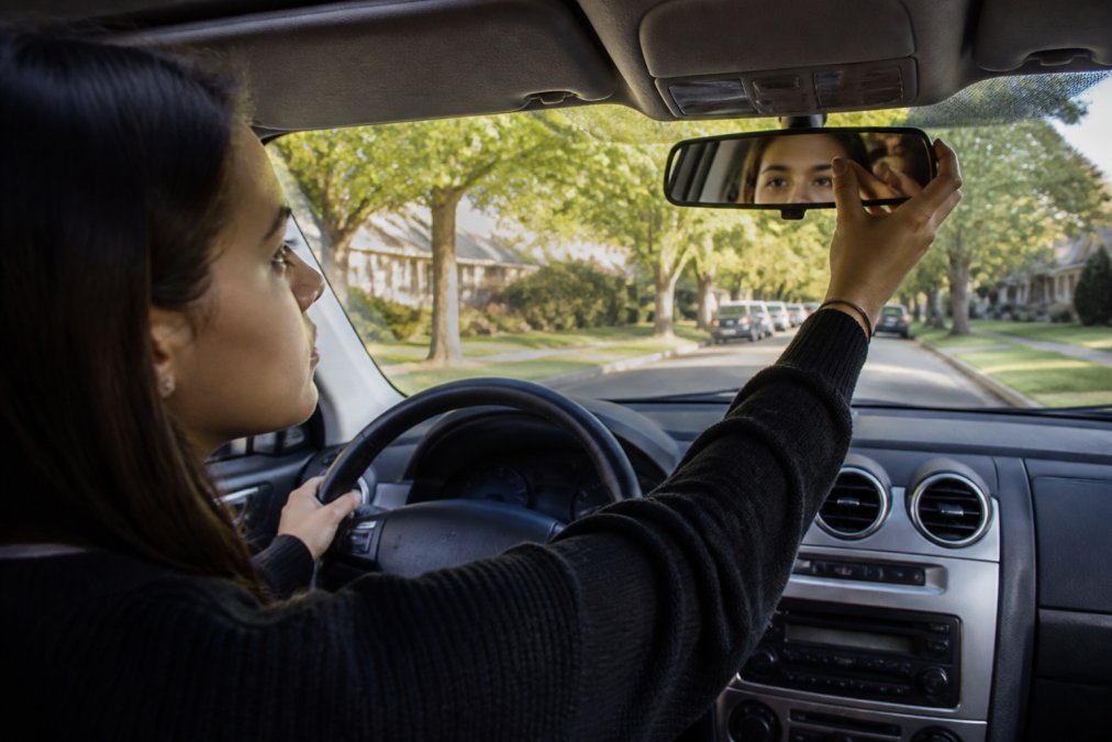 A calm, focused young adult sitting in the driver's seat of a car, hands on the wheel, adjusting the rearview mirror before starting a driving test, with a sunny suburban street visible through the windshield