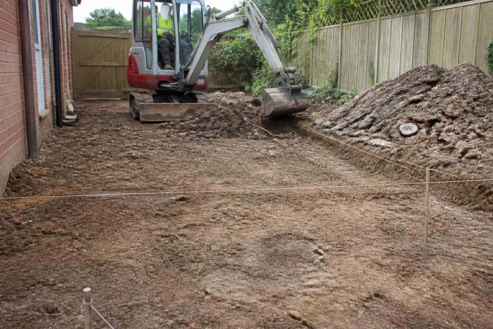 A driveway area being excavated with a small mechanical digger, showing exposed soil and removed rubble piled to the side, with groundwork markers and string lines visible across the prepared surface