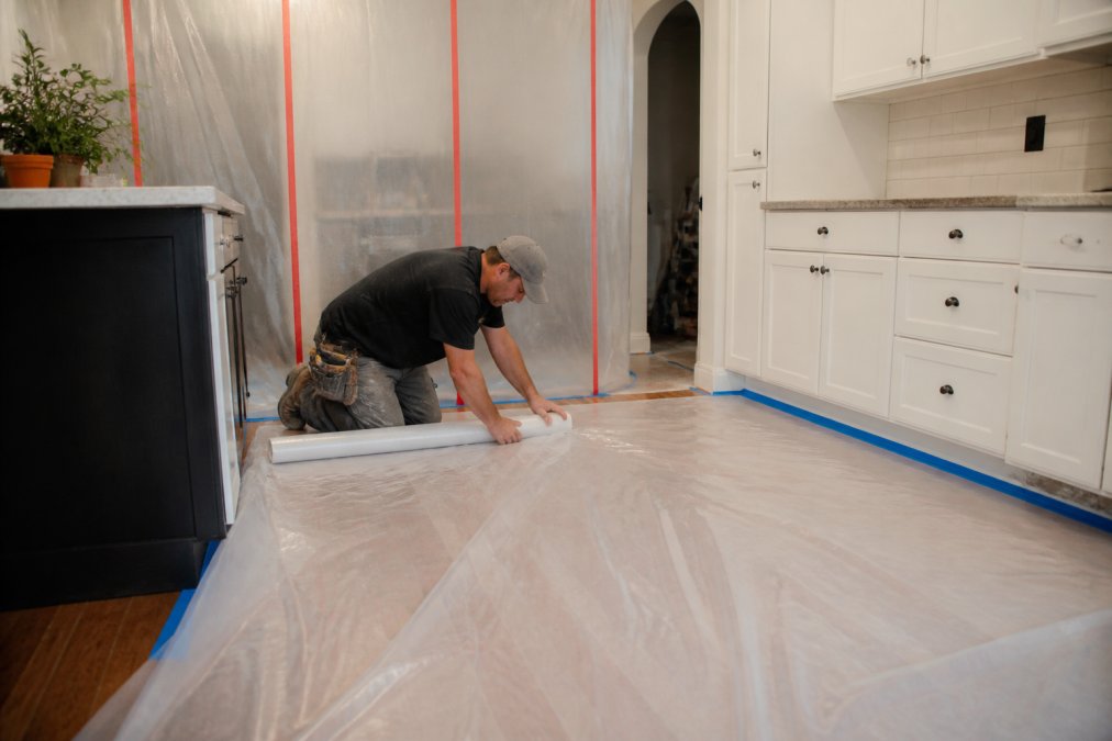 Construction workers laying plastic sheeting and protective coverings across hardwood flooring in a kitchen, taping edges to walls, with dust containment barriers visible in background