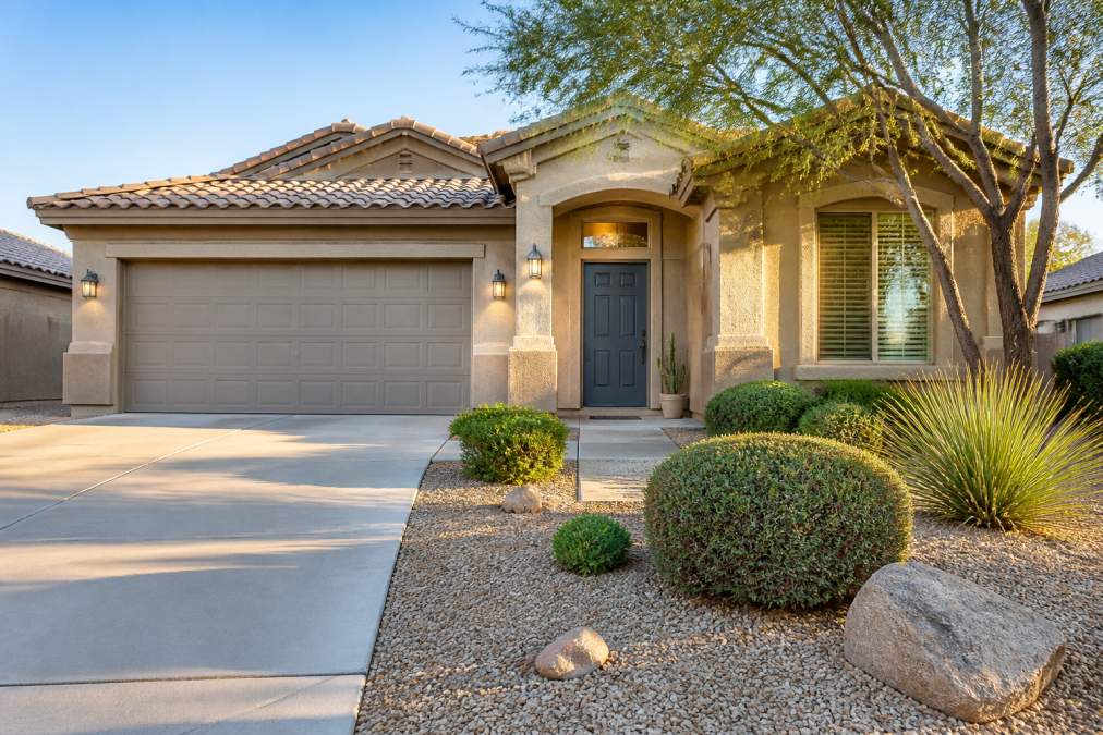 A well-maintained single-story home with neatly trimmed desert landscaping, a clean concrete driveway, and a freshly painted front door photographed in warm early morning light with soft shadows across the facade