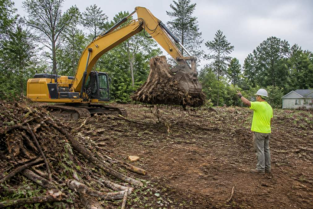 An excavator lifts a large tree stump to prepare land while a worker directs the debris removal on a residential lot.
