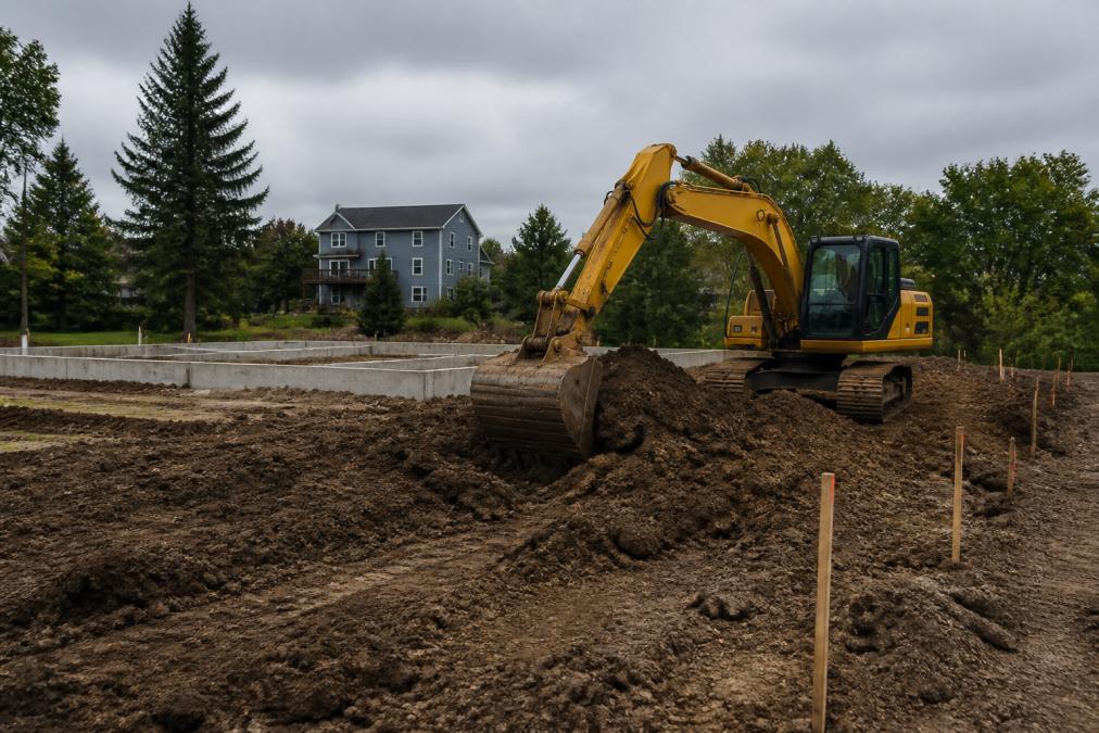 An excavator moving soil on a residential lot with a house foundation outline in the background, trees and wooden stakes visible, overcast Michigan sky