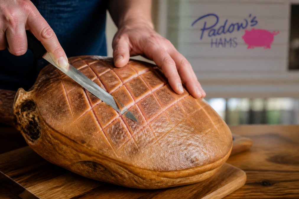 Close-up of hands scoring the rind of a salt-cured ham with a sharp knife, showing the diamond pattern being carved into the surface