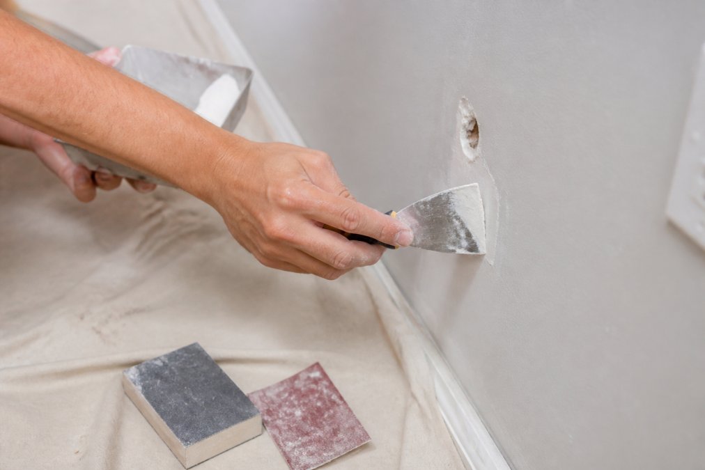 Hands using a putty knife to apply joint compound to prepare walls for painting near a sanding block.