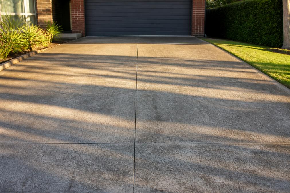 Cleared residential driveway with no vehicles or obstacles, showing bare concrete surface ready for pressure washing with morning sunlight casting long shadows across the clean pavement