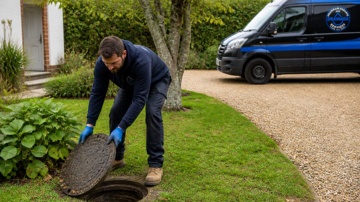 Homeowner lifting a heavy manhole cover in an East Sussex garden to prepare for a professional drainage inspection.