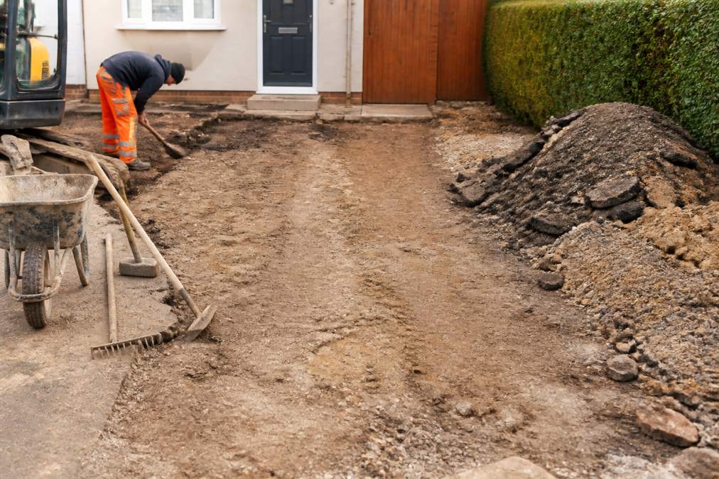 Professional removing old gravel and debris from a residential driveway area, revealing a compacted soil base, with piles of cleared material stacked neatly to the side
