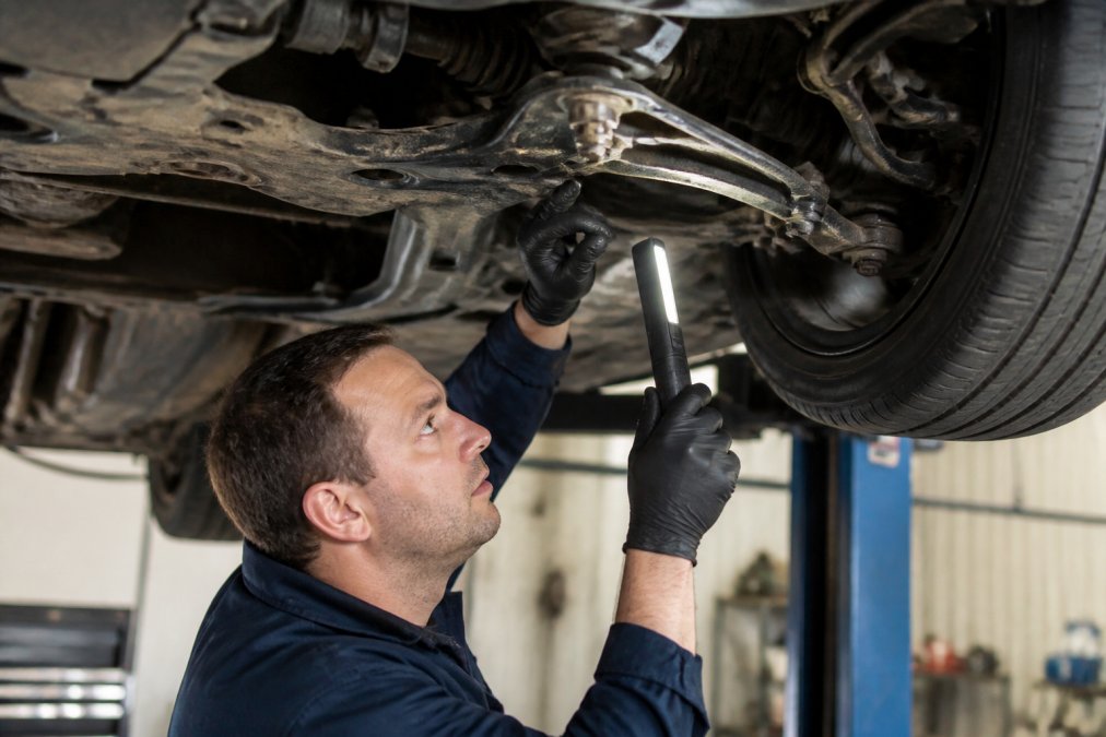 Mechanic performing a detailed inspection of a vehicle's undercarriage, checking suspension components and brakes with a torchlight in a service bay