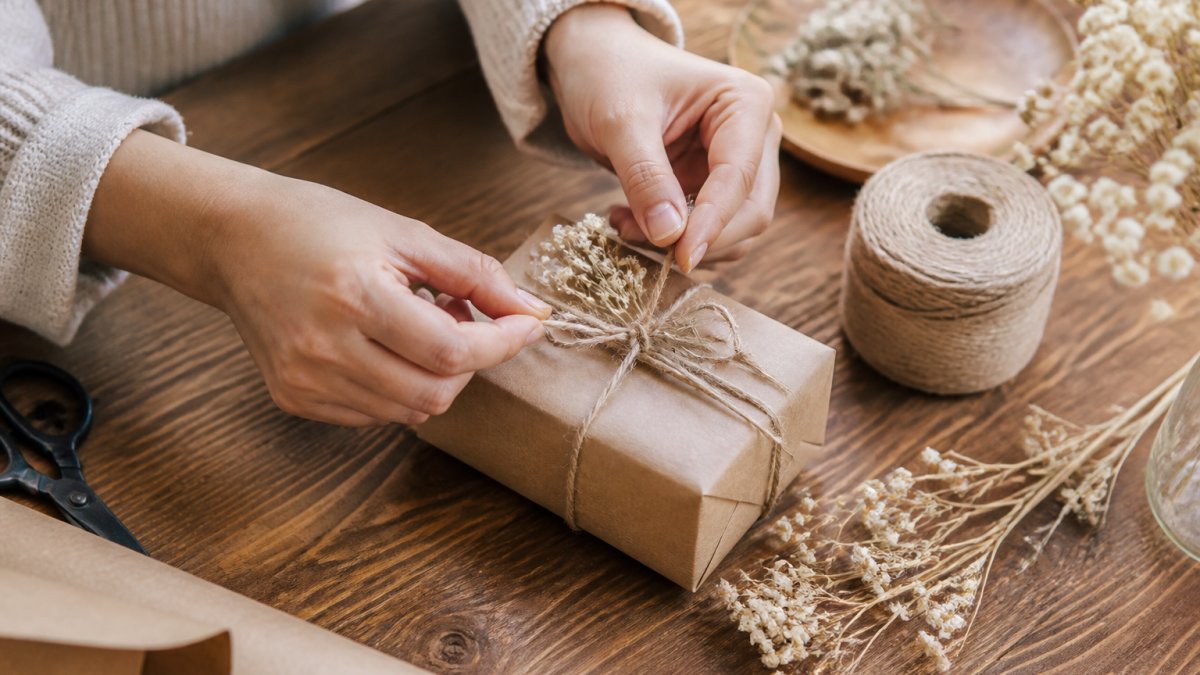 Hands wrapping a small personalized gift box with kraft paper, twine, and a handwritten note tag on a wooden table with dried flowers nearby