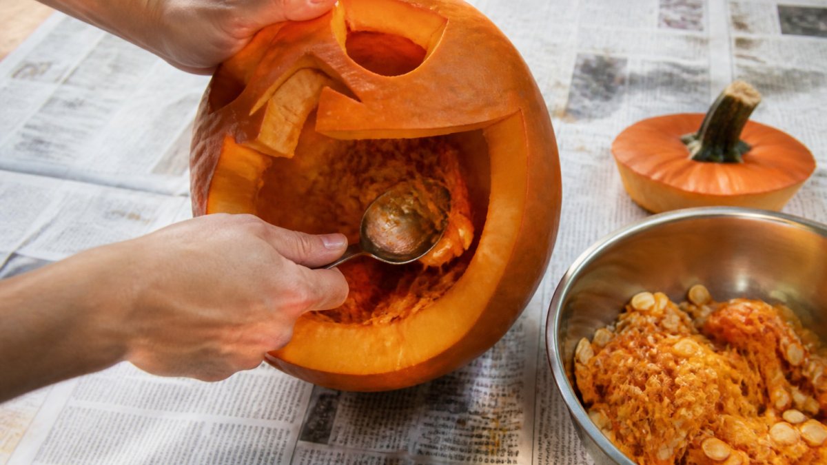 Close-up of hands scooping seeds and stringy pulp from inside a carved pumpkin on a newspaper-covered table, with a metal spoon and bowl nearby
