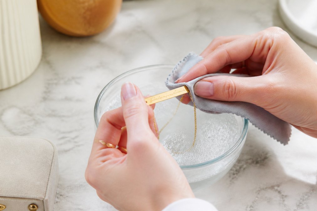 Close-up of hands gently cleaning an engraved handwritten necklace with a soft microfiber cloth and mild soapy water, showing careful attention to delicate script details in natural lighting
