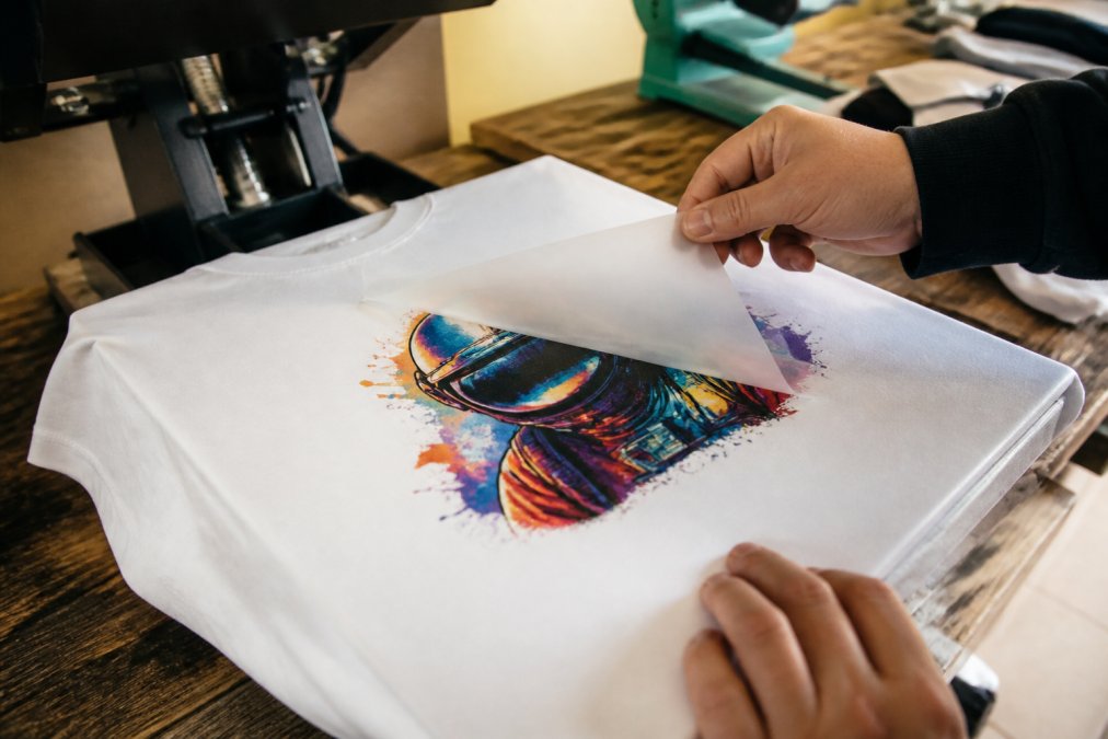 Hands carefully peeling back the transparent carrier film from a freshly pressed custom graphic on a white t-shirt laid flat on a heat press platen, the vibrant full-color design fully transferred to the fabric, warm workshop lighting overhead