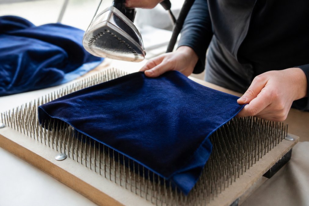 Close-up of hands placing a piece of deep blue velvet face-down onto a needle board, with a steam iron hovering just above the fabric surface in a bright sewing studio
