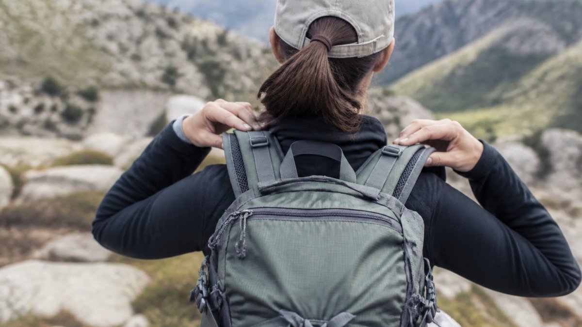 Hiker from behind adjusting padded shoulder straps on a backpack positioned high on back, hands positioning straps snug against shoulders in natural outdoor lighting