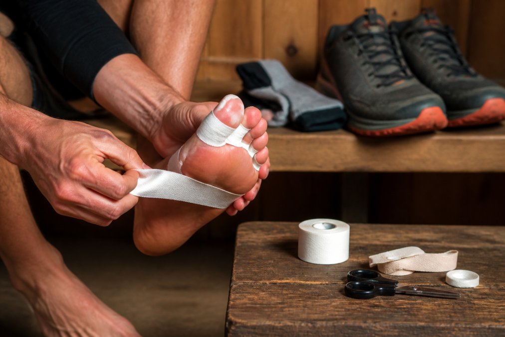 Hands carefully applying white athletic tape to a runner's toes and the ball of the foot before a trail run, trail shoes and folded performance socks resting on a wooden bench in the background, warm indoor lighting
