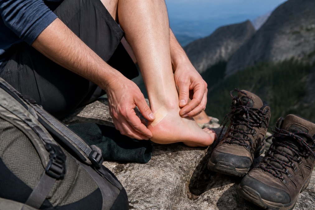 Close-up of a hiker sitting on a trailside rock, applying a blister patch to their heel, with hiking boots and backpack visible nearby
