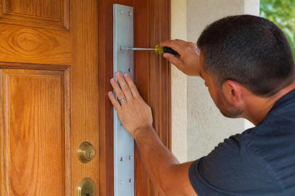 Close-up of a homeowner's hands installing a galvanized steel door reinforcement plate on a wooden front door frame, with a screwdriver and hardware nearby