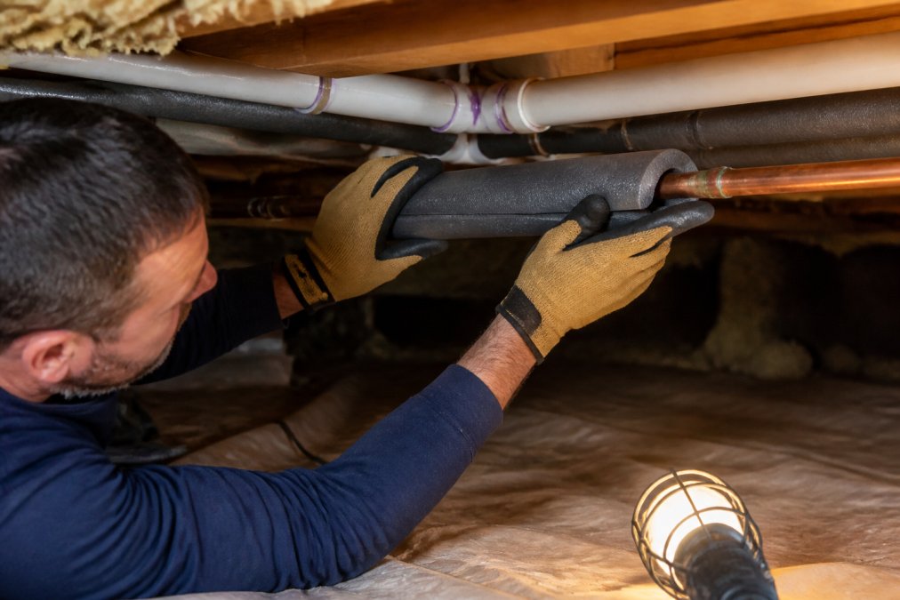 A homeowner in a crawl space installing foam pipe insulation on exposed copper water pipes, wearing work gloves and pressing a slit foam cover into place under a single work light
