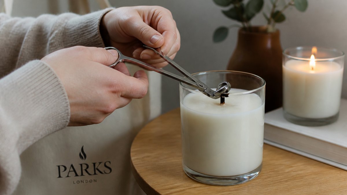 Close-up of hands using wick trimmers to cut a blackened candle wick to about 5mm above the wax surface, with a lit candle softly blurred in the background on a wooden side table