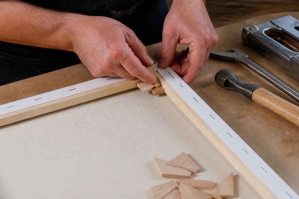 Close-up of hands carefully adjusting corner keys on the back of a stretched canvas frame, fingers positioned to demonstrate the proper tensioning technique with wooden stretcher bars visible
