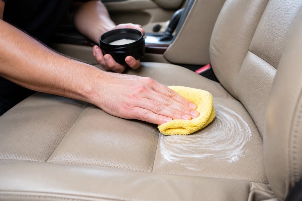 Close-up of hands applying leather conditioner to car seat with microfiber cloth, showing proper circular motion technique and even product distribution across the leather surface