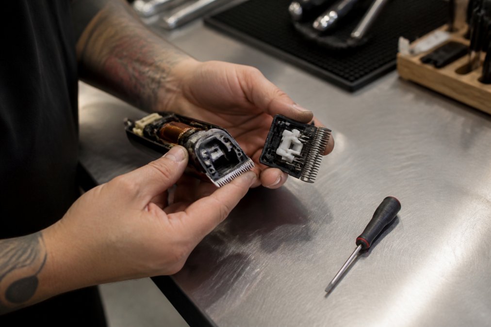A barber's hands holding open clippers with the blade set removed, inspecting blade alignment under bright overhead shop lighting on a stainless steel work surface, with a small flathead screwdriver resting nearby
