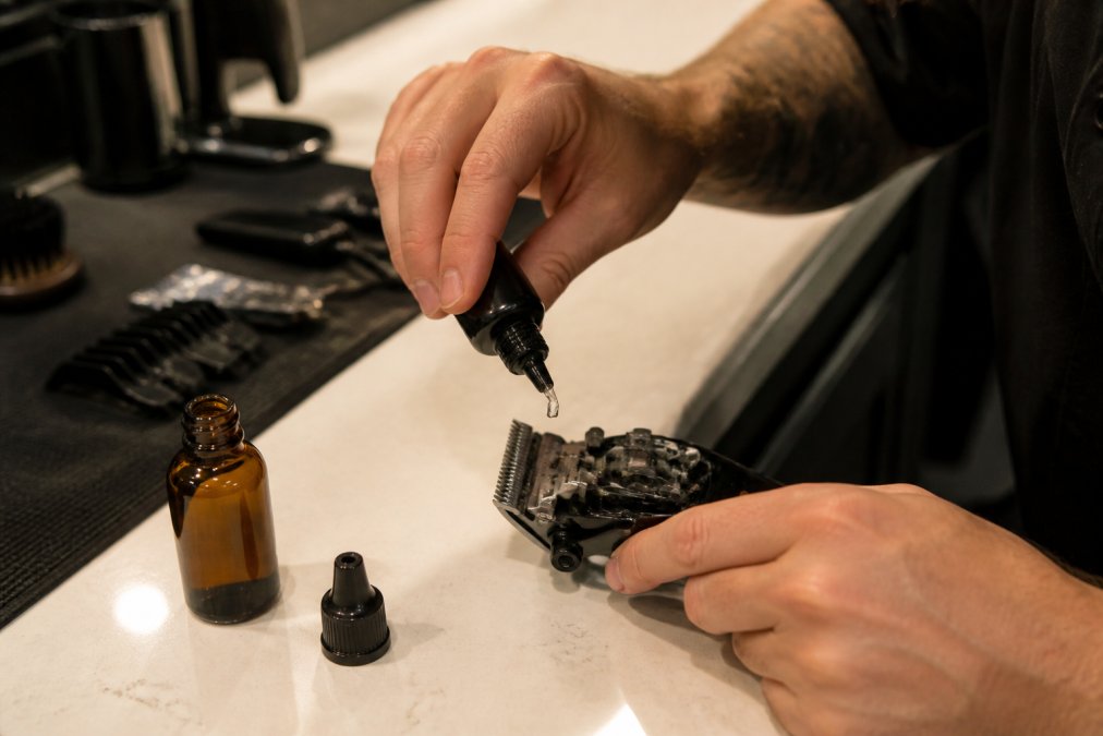 Close-up of a barber's hands applying small drops of clipper oil onto open clipper blades resting on a clean white barbershop counter, with a small amber oil bottle visible in the frame under warm overhead lighting