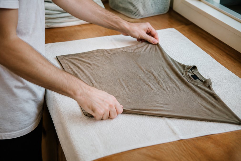 Hands gently stretching a damp cotton garment laid flat on a clean white towel on a timber laundry bench, soft indoor lighting