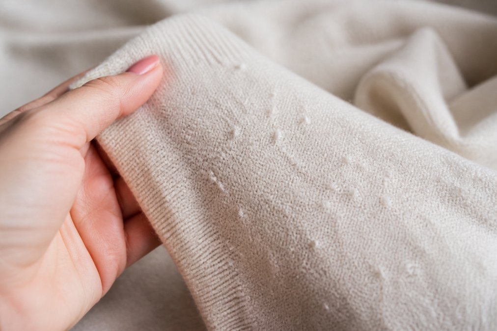 Close-up of a woman's hand holding a light-colored sweater sleeve, showing small fabric pills on the fabric against a soft neutral background