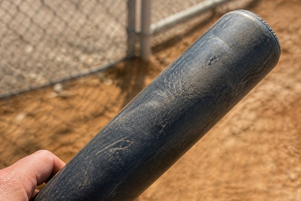 Close-up of composite bat barrel showing spider webbing cracks and hairline fractures under natural light, with the bat held at an angle to clearly display damage patterns
