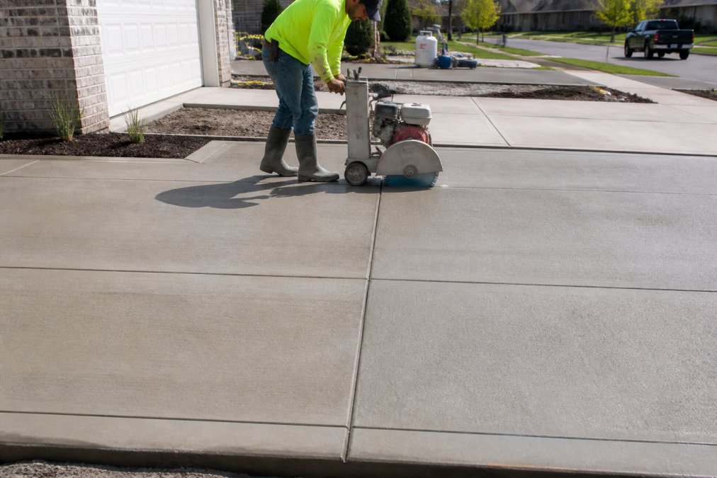 Professional concrete contractor using a concrete saw to cut control joints into a freshly poured driveway, showing the precise spacing pattern and straight lines being cut into gray concrete