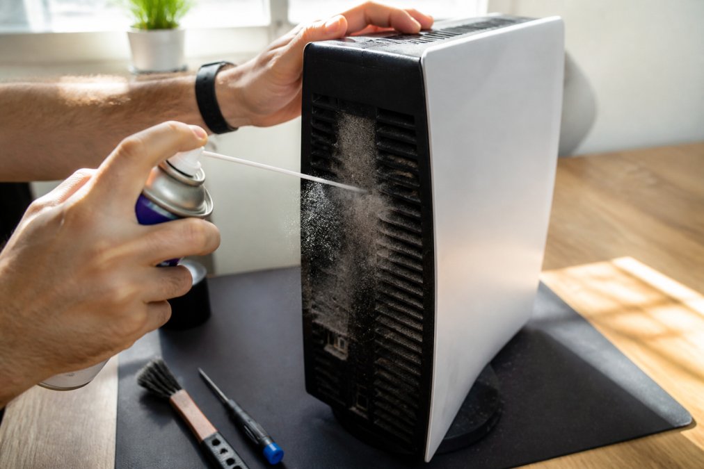 Hands carefully removing dust from console vents using compressed air can, with visible dust particles floating away from the device in bright natural lighting
