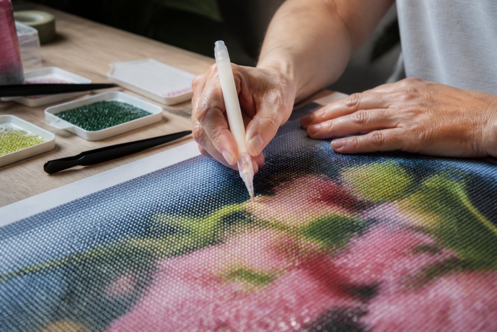 Close-up of hands using a specialized drill pen to place a diamond drill onto a sticky canvas, with a light pad glowing underneath
