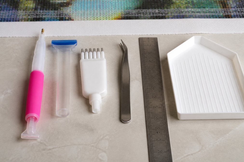 Flat-lay arrangement of diamond painting tools including ergonomic drill pen with comfortable grip, diamond painting straightening tool, multiplacer with multiple drill slots, stainless steel tweezers, and metal ruler positioned next to an open drill tray on a craft work surface