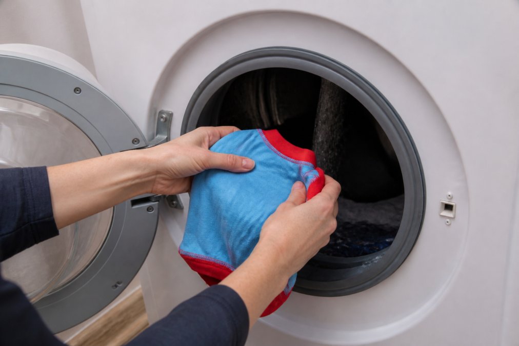 Close-up of hands placing a colorful dog t-shirt into a washing machine with cold water running, showing proper loading technique with the garment turned inside out