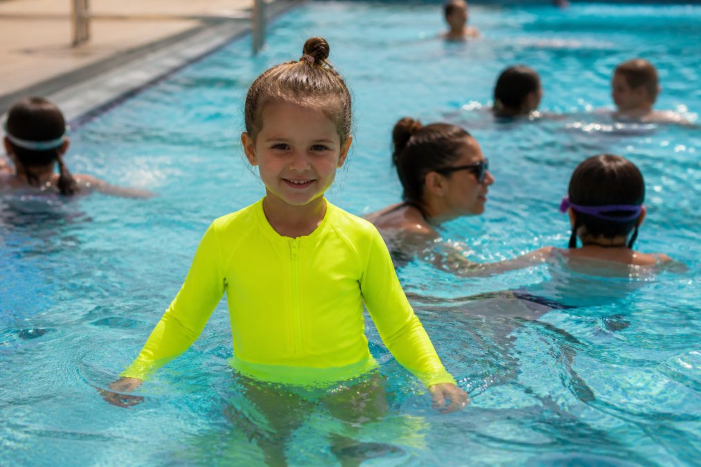 Young child wearing a bright neon-colored high-visibility swimsuit standing in a public pool, distinctly visible against the water and other swimmers