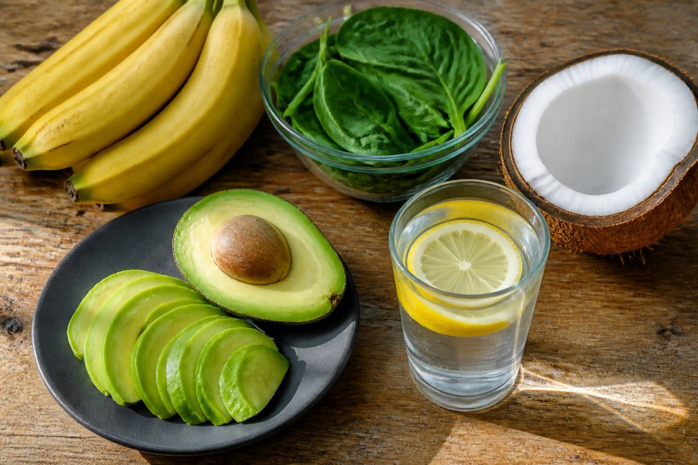 Overhead shot of a rustic wooden table with bananas, sliced avocado, a bowl of leafy greens, a halved coconut, and a glass of lemon water in soft daylight