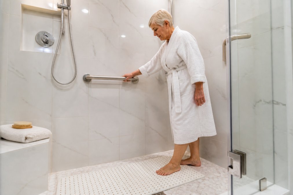 An older adult gripping a sturdy grab bar inside a walk-in shower, with a non-slip mat on the floor and bright overhead lighting illuminating the white tile walls