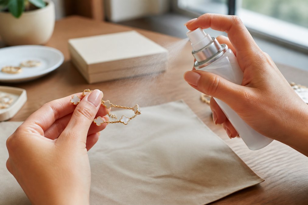 Close-up of hands carefully applying clear jewelry sealant spray to a delicate gold-toned bracelet, held at proper distance over a protective surface